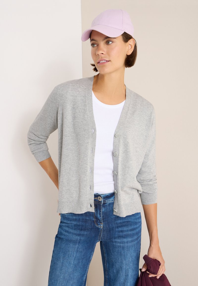 Cardigan grigio chiaro con scollatura a V e bottoni frontali, abbinato a una t-shirt bianca e jeans blu. Tiene un oggetto bordeaux con una mano.