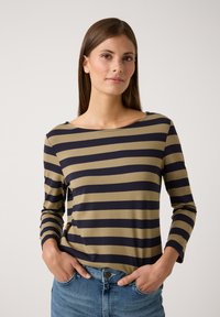 Langärmliges Shirt in olivgrün mit navyblauen Streifen, mit einem Bootsausschnitt und weichem Stoff. Das Model pose mit den Händen in den Taschen.