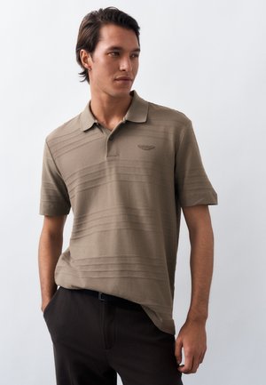 Mężczyzna ubrany w brązową, teksturowaną koszulkę polo z krótkim rękawem i kołnierzykiem, w parze z czarnymi spodniami, stojący z jedną ręką w kieszeni.