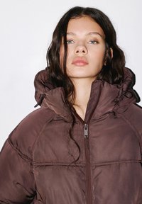 Braune Pufferjacke mit hohem Kragen und verstellbarem Kapuze. Verfügt über einen Frontreißverschluss und eine gestepptes Muster. Glattes, glänzendes Material.