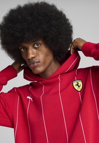 Rød hættejakke med hvide lodrette striber, udstyret med et Ferrari-logo patch. Lavet af blødt stof, den har en hætte med snoretræk og ribbede manchetter.