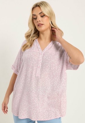 Frau mit blondem Haar, die eine lockere, kurzärmelige rosa Bluse mit floralem Muster und helle blaue Jeans trägt, wobei sie mit einer Hand ihre Haare berührt.