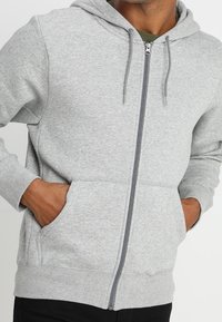 Felpa grigia chiara con zip, realizzata in tessuto morbido, dotata di cappuccio, tasche frontali e lacci. Design semplice con vestibilità standard.