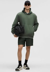 Sudadera verde oliva con gran bolsillo frontal, combinada con pantalones cortos de malla verde oscuro. Accesorio: mochila negra; calzado: zapatillas gris y negro.