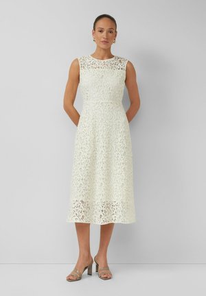 Kobieta w białej, koronkowej sukience midi bez rękawów i beżowych sandałach na obcasie, stojąca na tle gładkiego, jasnoszarego tła.