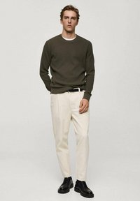 Khakifarbener gerippten Pullover, cremfarbene Hose, schwarzer Gürtel und schwarze Schnürschuhe. Das Model steht in einer neutralen Umgebung mit den Händen in den Taschen.