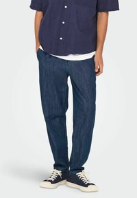 Pantaloni din denim de culoare bleu închis, cu croială dreaptă, având o textură subtilă și un design clasic cu cinci buzunare, asortați cu tenisi casual.
