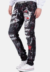 Jeans in denim nero con dettagli strappati, vernice rossa e bianca spruzzata, e disegni grafici di teschi, con una vestibilità slim e hardware accentuato.