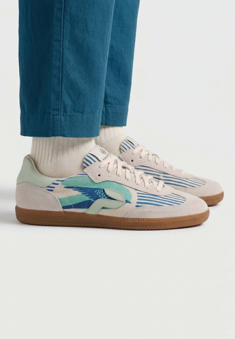 Pieds chaussés de baskets beiges et vertes avec des motifs d'oiseau bleu et de rayures, assortis à des chaussettes blanches et un pantalon bleu sarcelle.