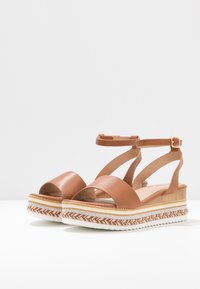 Sandales compensées en cuir marron avec des sangles croisées à la cheville, des détails en jute tressé et une semelle en caoutchouc blanc. Fort contraste de textures avec le cuir lisse.