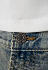Gros plan sur le bouton d'un jean en denim bleu fermé sur un tissu blanc cousu, montrant les détails de la texture et des coutures.