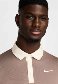 Nike polo krekls gaiši brūnā krāsā ar krēmkrāsas apkakli un pogu rindu, ar balto logotipu uz krūtīm. Gluda tekstūra.