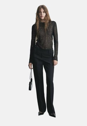 Schwarzes, transparentes Oberteil mit langen Ärmeln und verstreuten Verzierungen, kombiniert mit maßgeschneiderten schwarzen Hosen. Das Model hält eine kleine schwarze Tasche.