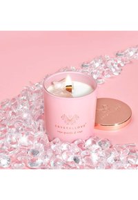 Różowa świeca w szklanym słoiku z złotym akcentem i białym woskiem, otoczona przez przezroczyste dekoracje przypominające kryształy na różowym tle.