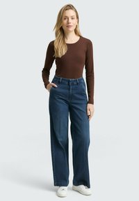 Braunes langärmliges Oberteil kombiniert mit hochgeschnittenen, weiten blauen Denim-Jeans. Das Model trägt weiße Sneakers und hat die Hände in den Taschen.