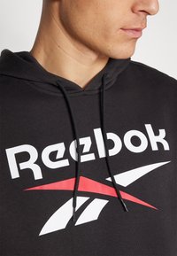 Crna kapuljača s kangaroo džepom, s odvažnim bijelim natpisom "Reebok" i crvenim akcentom loga. Vidljivi su vezice i mekani materijal.