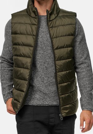 Hombre con chaleco acolchado color verde oliva sin cremallera sobre suéter gris de punto y pantalones negros, con la mano descansando cerca del bolsillo.