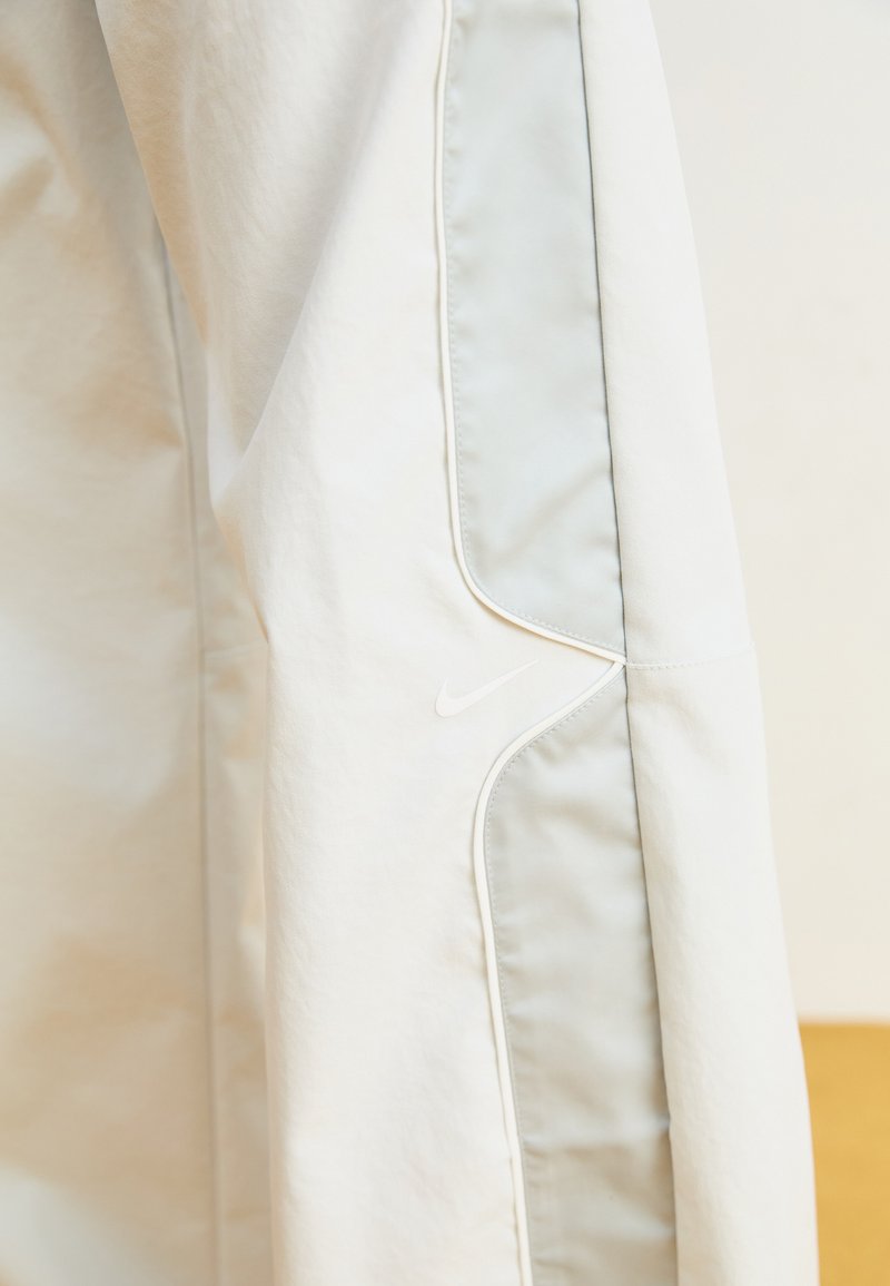 Hellegraue Sportjacke mit weißem Nike-Logo, glatter Textur, kontrastierendem Paneeldesign und minimalen Nähedetails.