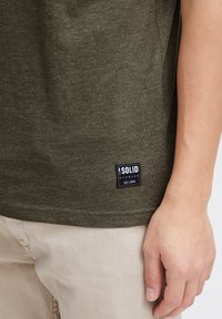 T-shirt vert olive en coton doux avec un col rond et une étiquette noire rectangulaire en bas à gauche, comportant un texte blanc.