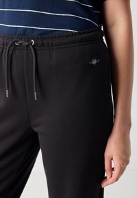 Svarte joggebukser med snor i midjen, med en liten brodert logo. Jevn tekstur, innsvingt passform og sidelommer.