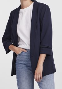 Marinblå blazer med avspänd passform, öppen framsida och trekvartsärmar, ihopparad med en enkel vit skjorta och ljusblå jeans.