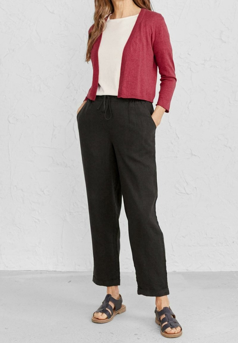 Femme portant un haut blanc, un cardigan rouge court, un pantalon noir avec cordon de serrage et des sandales noires à bouts ouverts, debout contre un mur blanc texturé.