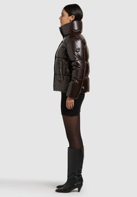 Brown puffer jacket with a high collar, shiny finish, zippered pockets, and logo detailing. Worn with a black dress and knee-high boots.