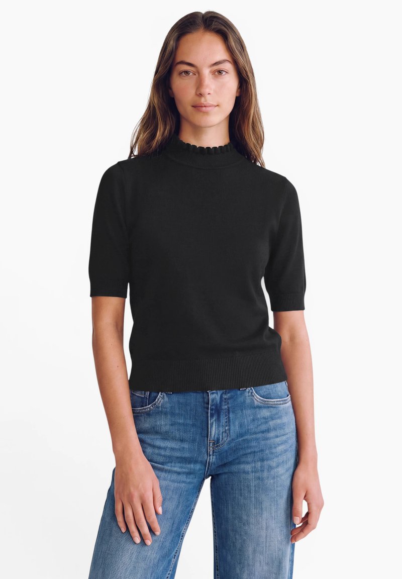 Femme portant un pull noir à manches courtes avec un col montant à volants et un jean en denim bleu sur fond blanc uni.