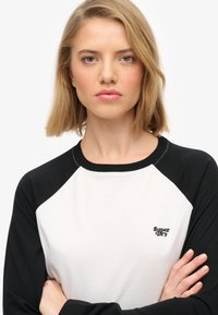 Biała koszulka baseballowa z czarnymi rękawami raglanowymi, z małym czarnym logo "Super Dry" na piersi. Wykonana z bawełny, o regularnym kroju.