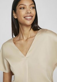 Bežinė, trumpomis rankovėmis palaidinė, pagaminta iš minkštos, lygios medžiagos, su V formos iškirpte ir minimaliais siuvimo akcentais.