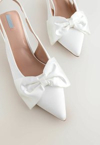 Valged satiiniteedega slingback kontsad, millel on terav nina ja suur pael ees. Siledat tekstuuri ja heledat pruunikat sisetald.