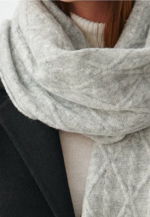 Femme portant une épaisse écharpe tricotée gris clair sur un pull blanc et un manteau noir, ne montrant que la poitrine et le cou.