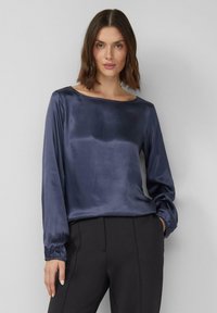 Donkerblauwe satijnen top met lange mouwen, een boothals en geplooide manchetten. Gladde textuur en een relaxte pasvorm. Gecombineerd met zwarte broek.