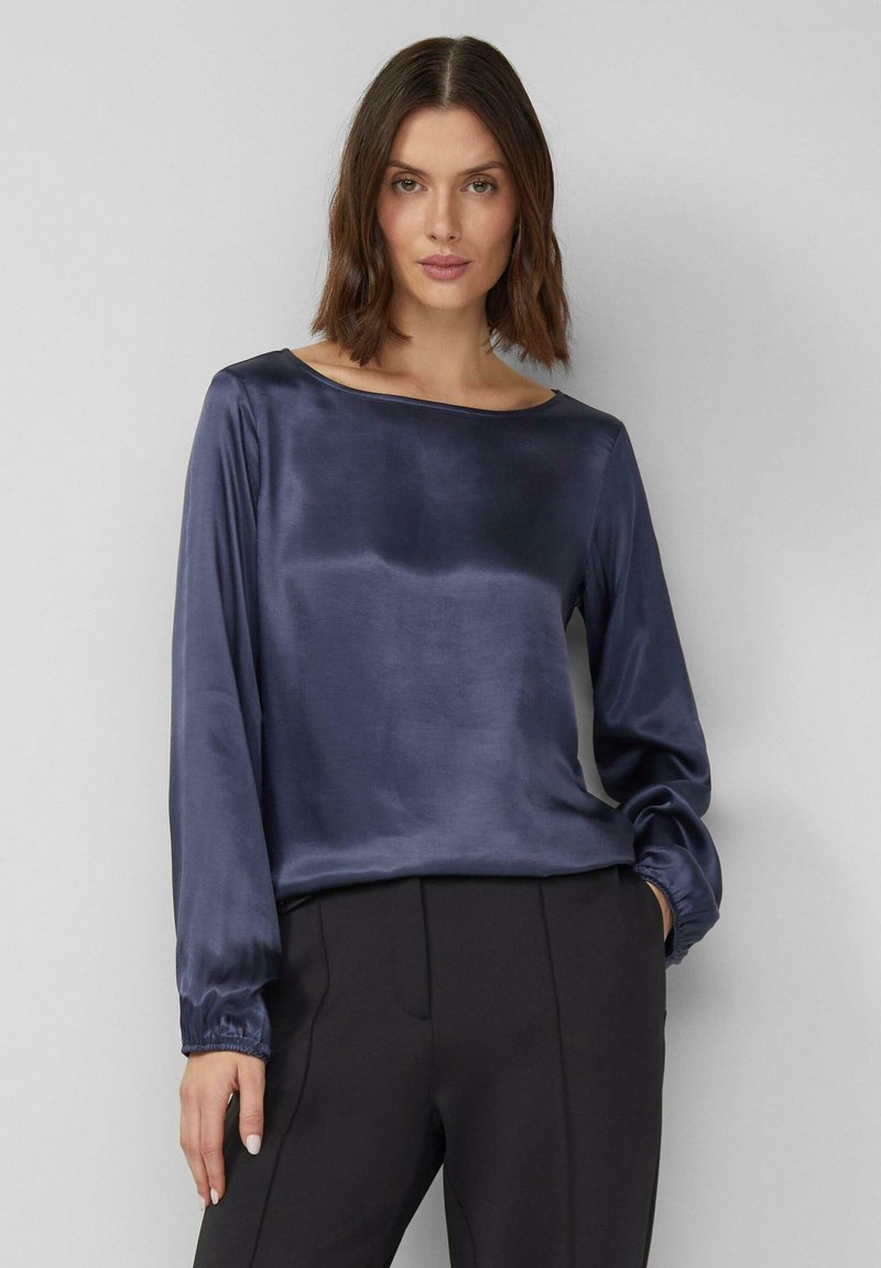 Donkerblauwe satijnen top met lange mouwen, een boothals en geplooide manchetten. Gladde textuur en een relaxte pasvorm. Gecombineerd met zwarte broek.