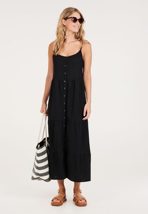 Schwarzes Maxikleid mit schichtiger Gestaltung, mit vorderen Knöpfen, Spaghetti-Trägern und einer fließenden Silhouette. Begleitet von einer gestreiften Tasche und Sandalen.