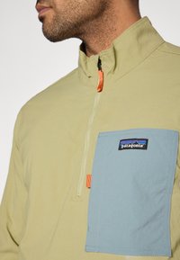 Ανοιχτοπράσινο πουλόβερ με φερμουάρ τέταρτου μήκους σε πορτοκαλί χρώμα, με τσέπη στο στήθος σε μπλε χρώμα που φέρει το λογότυπο της Patagonia. Υφή λεία.