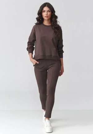 Kobieta o długich ciemnych włosach idzie do przodu, ubrana w brązową bluzkę z długim rękawem, dopasowane spodnie i białe trampki na jednolitym szarym tle.