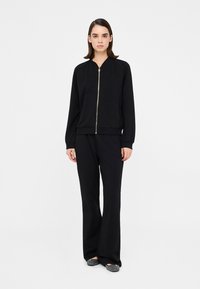 Čierna bomber bunda na zips s rebrovan�ými manžetami, v kombinácii so širokými nohavicami. Obe sú vyrobené z hladkej textílie, majú voľný strih a minimalistický dizajn.