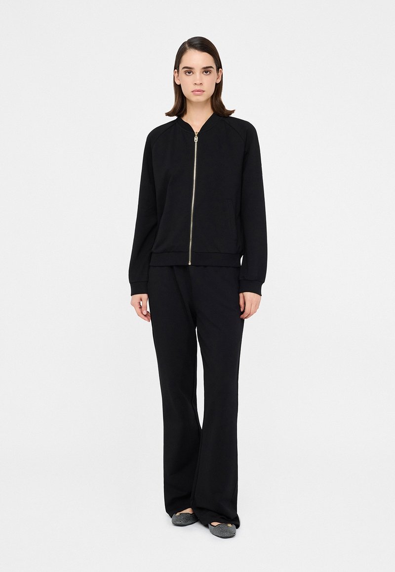 Čierna bomber bunda na zips s rebrovanými manžetami, v kombinácii so širokými nohavicami. Obe sú vyrobené z hladkej textílie, majú voľný strih a minimalistický dizajn.