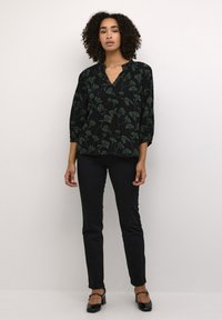 Blouse noire avec motif de feuilles vertes, décolleté en V, manches trois-quarts. Associée à un pantalon noir ajusté et des chaussures noires à bout fermé.