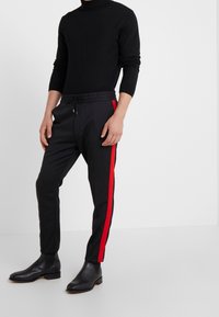 Maglione nero a collo alto, pantaloni sportivi neri slim-fit con una striscia laterale rossa e stivaletti neri in pelle alla caviglia. Vita elastica sui pantaloni.
