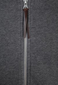 Indumento grigio con cerniera metallica e tirante in pelle marrone. Tessuto morbido con trama a coste in tutta la superficie, caratterizzato da un design con collo alto.