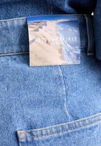 Denimjeans i blå nyans med en tygetikett som visar en strandscen och texten "Fête Impériale Paris." Sömmen är synlig.