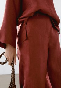 Osoba w luźnych czerwonych spodniach i dopasowanej bluzce, trzymająca ciemno-brązową torebkę z koralikami przy boku.