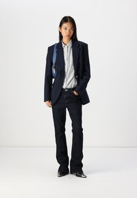 Blazer navy, chemise rayée grise, jean en denim foncé et chaussures noires brillantes ; présente une bandoulière bleue et une coupe élégante.