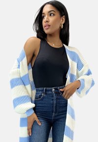 Langer, offener Cardigan aus Strickstoff mit abwechselnden blauen und weißen Streifen. Getragen über einem schwarzen Tanktop und hochgeschnittenen Jeans.