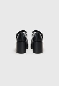 Black platform shoes with a smooth leather upper, back strap, and chunky heel. Notable round cutouts along the sides for ventilation.