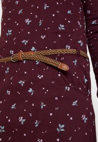 Bordeauxfarbenes Kleid mit floralem Muster in Hellblau und Pink, ausgestattet mit einem geflochtenen, braunen Gürtel und einer Vordertasche für zusätzliche Details.