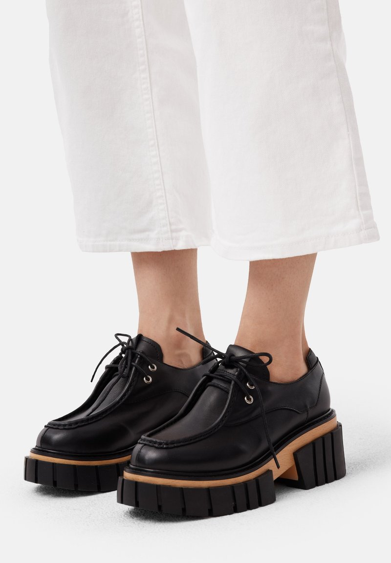Zapatos de cuero negro con cordones, suela exterior de goma negra gruesa y acento de madera, con puntera redondeada y detalles de costura mínimos.