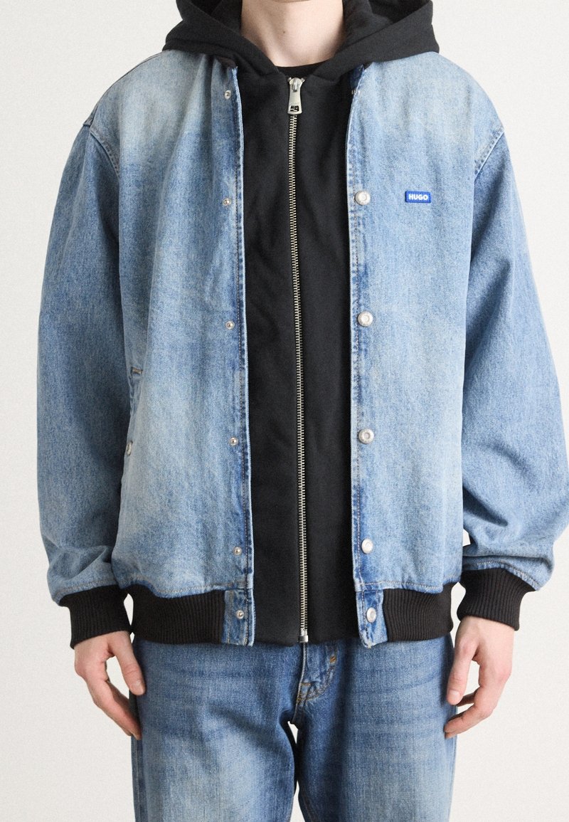 Denim jacket in light blue with black ribbed cuffs, featuring snap buttons and a small blue logo. Layered over a black zip-up hoodie.